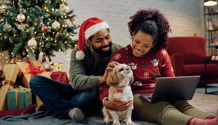 a man and woman sitting by a Christmas tree with a laptop and petting their dog